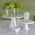 Three white pedestal cake stands arranged on a marble countertop, with two clear drinking glasses on the stands and two green hydrangea arrangements in tall clear glasses in the background.