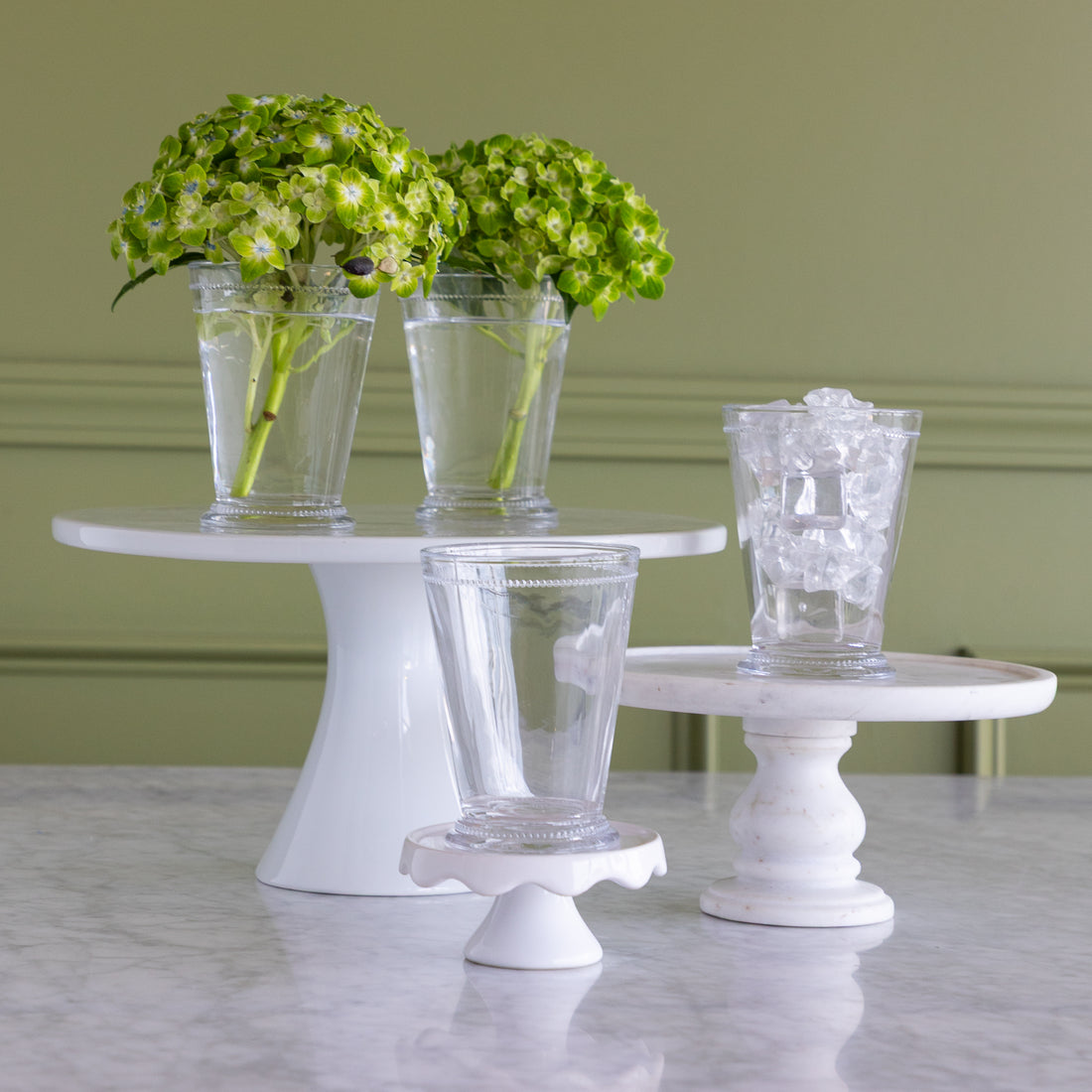 Three clear glass drinking glasses displayed on white pedestal stands against a sage green wall and marble tabletop. Two stands hold green hydrangea arrangements in tall glasses, and one stand holds a shorter clear glass filled with ice.