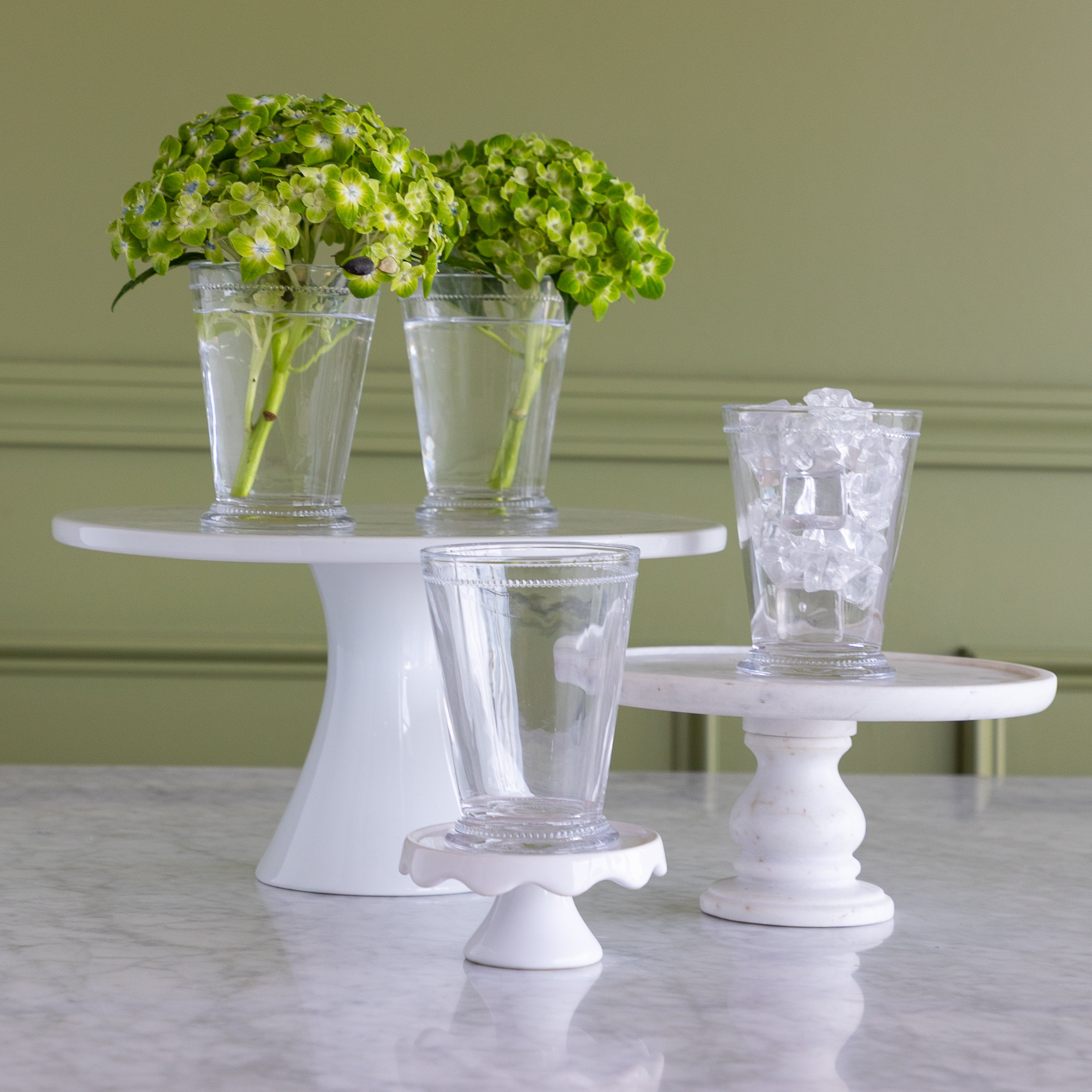 Three clear glass drinking glasses displayed on white pedestal stands against a sage green wall and marble tabletop. Two stands hold green hydrangea arrangements in tall glasses, and one stand holds a shorter clear glass filled with ice.