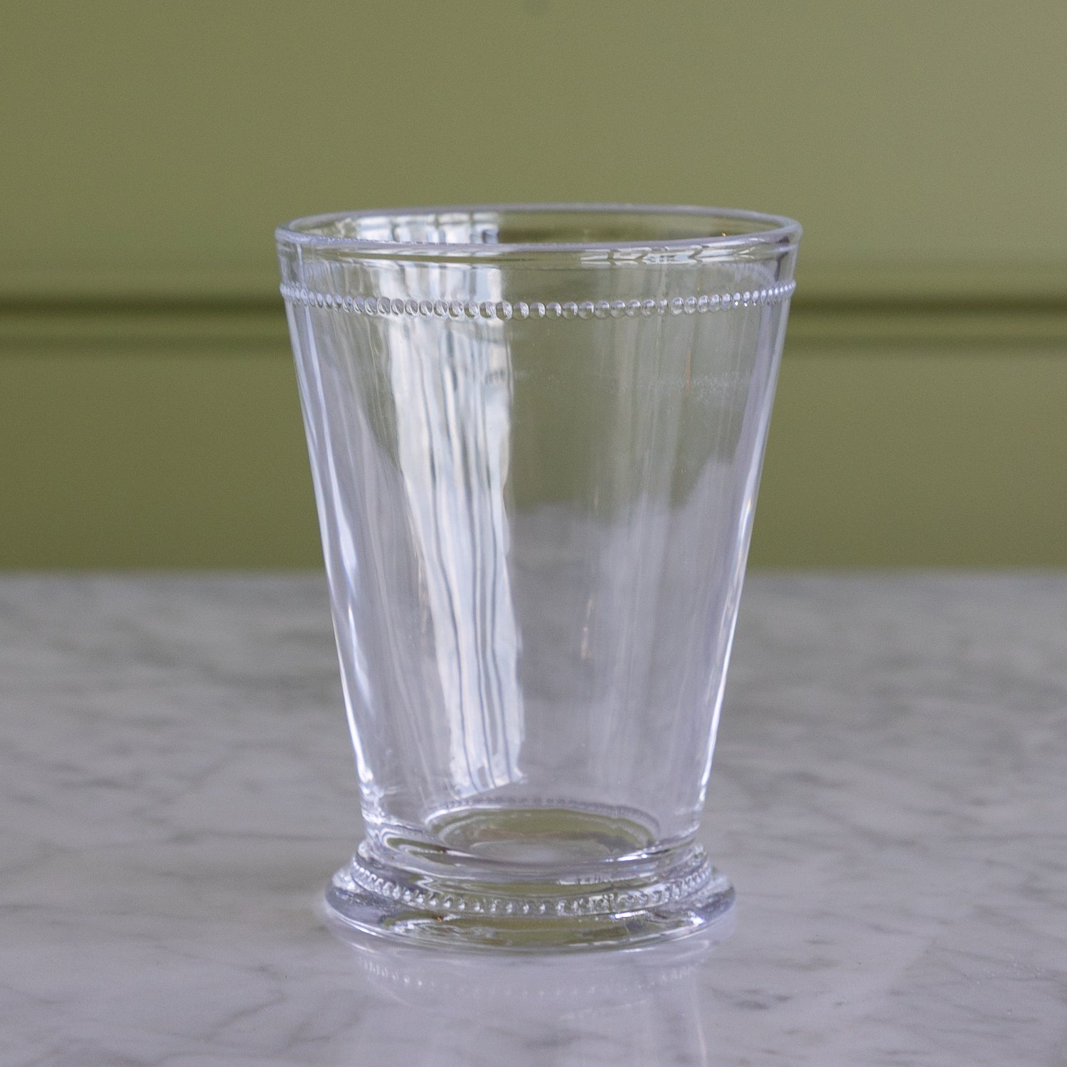 A clear glass tumbler with a decorative beaded rim and base, standing on a white marble surface against a muted olive green wall.