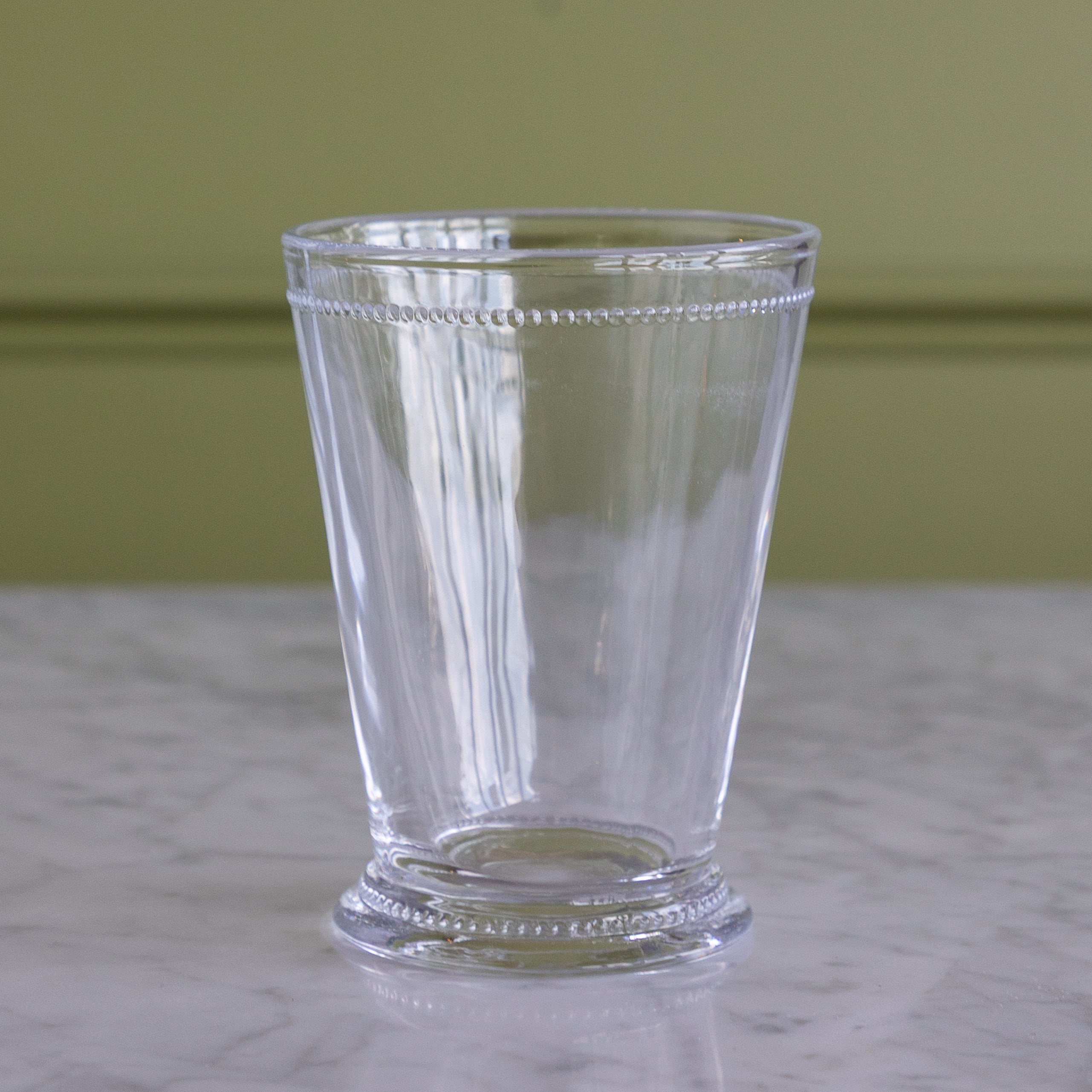 A clear glass tumbler with a decorative beaded rim and base, standing on a white marble surface against a muted olive green wall.