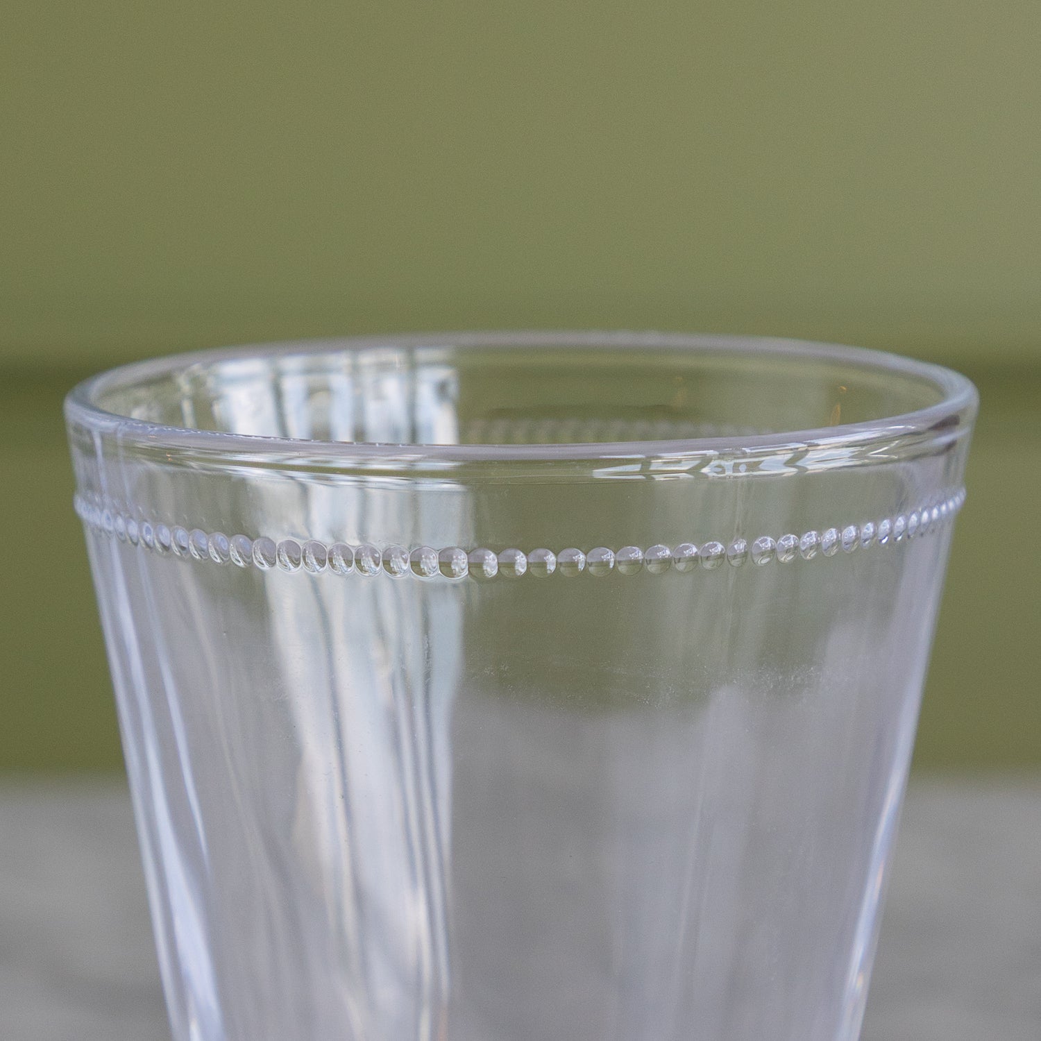 A close-up of a clear glass tumbler with a decorative beaded rim, set against a soft olive green background.