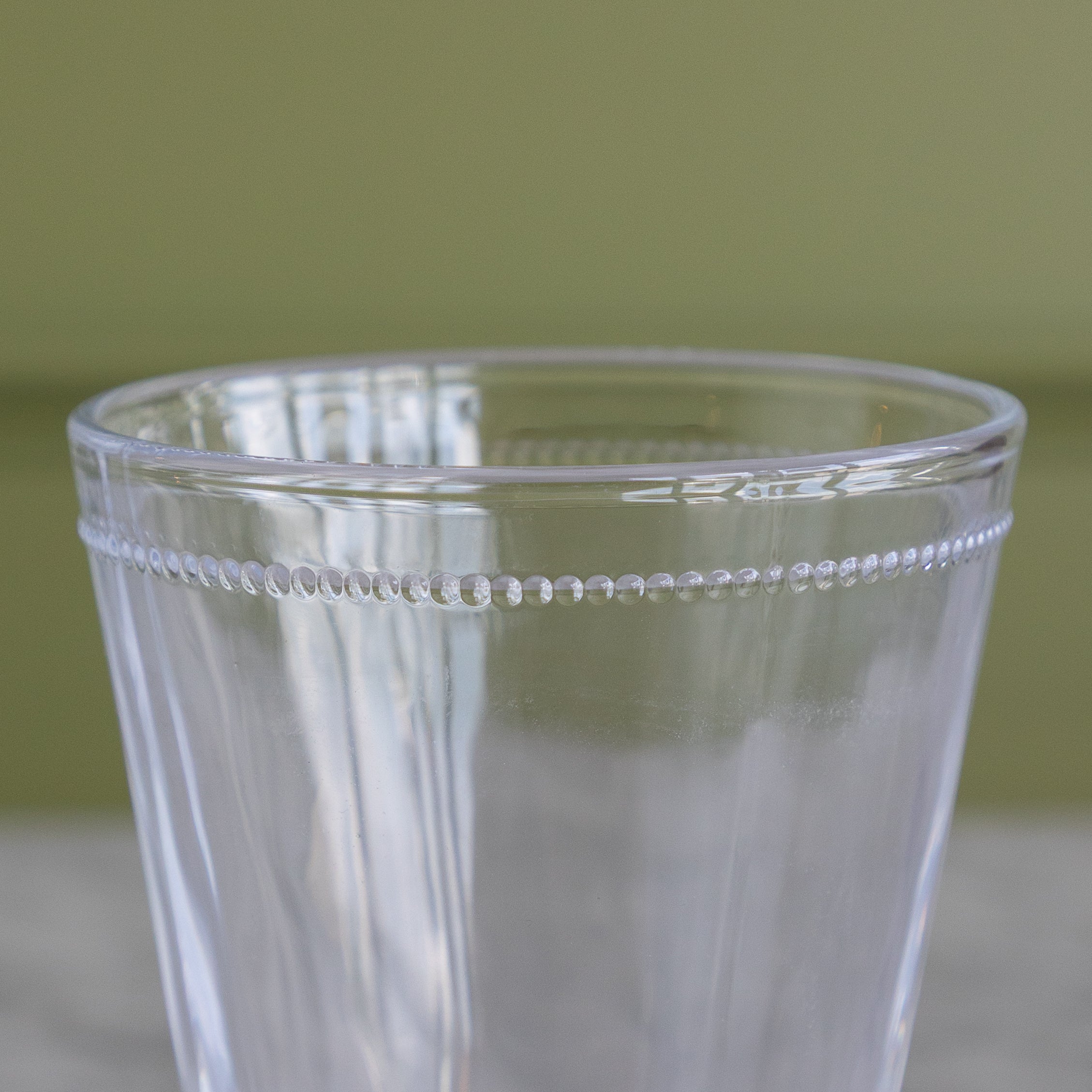 A close-up of a clear glass tumbler with a decorative beaded rim, set against a soft olive green background.