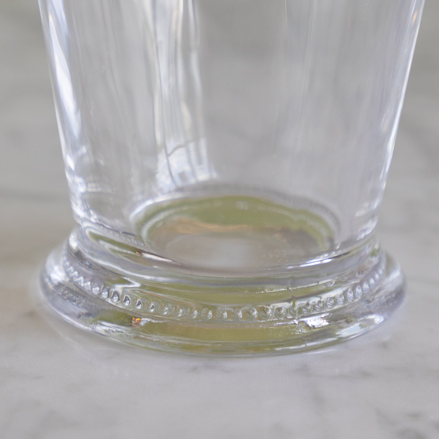 A close-up of a clear glass drinking cup with a decorative beaded base, resting on a light marble surface.