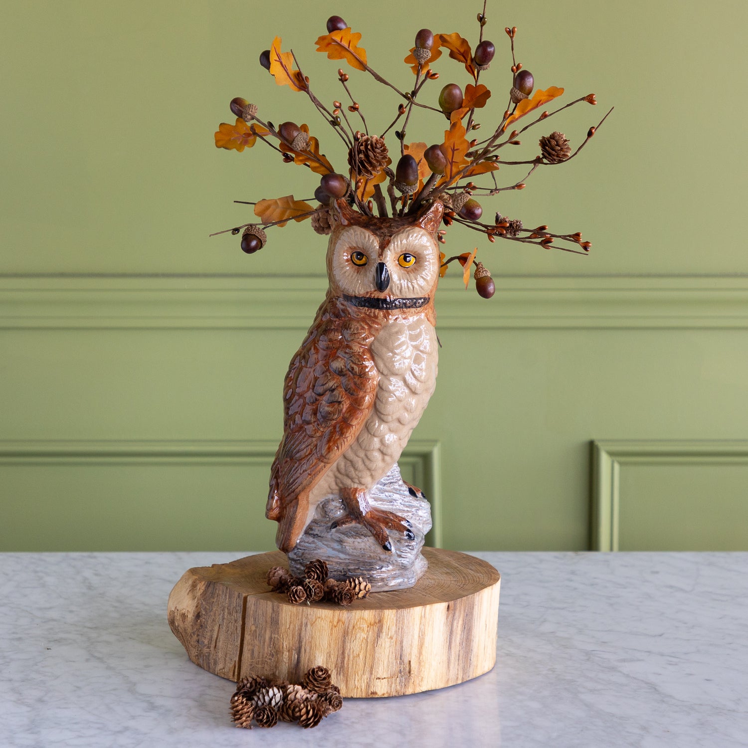 A decorative owl figurine perched on a faux log base, with a branch arrangement of orange leaves, acorns, and pinecones extending from its head, displayed on a marble surface against a green paneled wall.