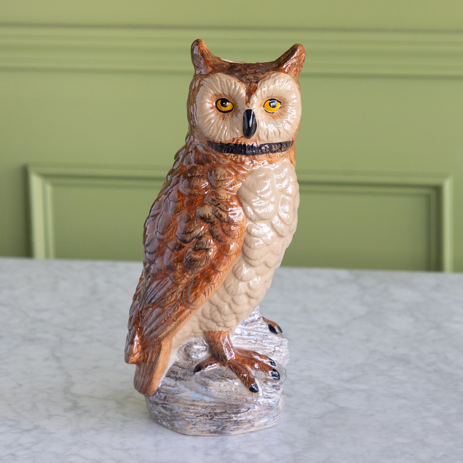 A ceramic owl figurine with brown and cream feathers, yellow eyes, and a black beak, standing on a textured base against a green paneled wall and white marble surface.