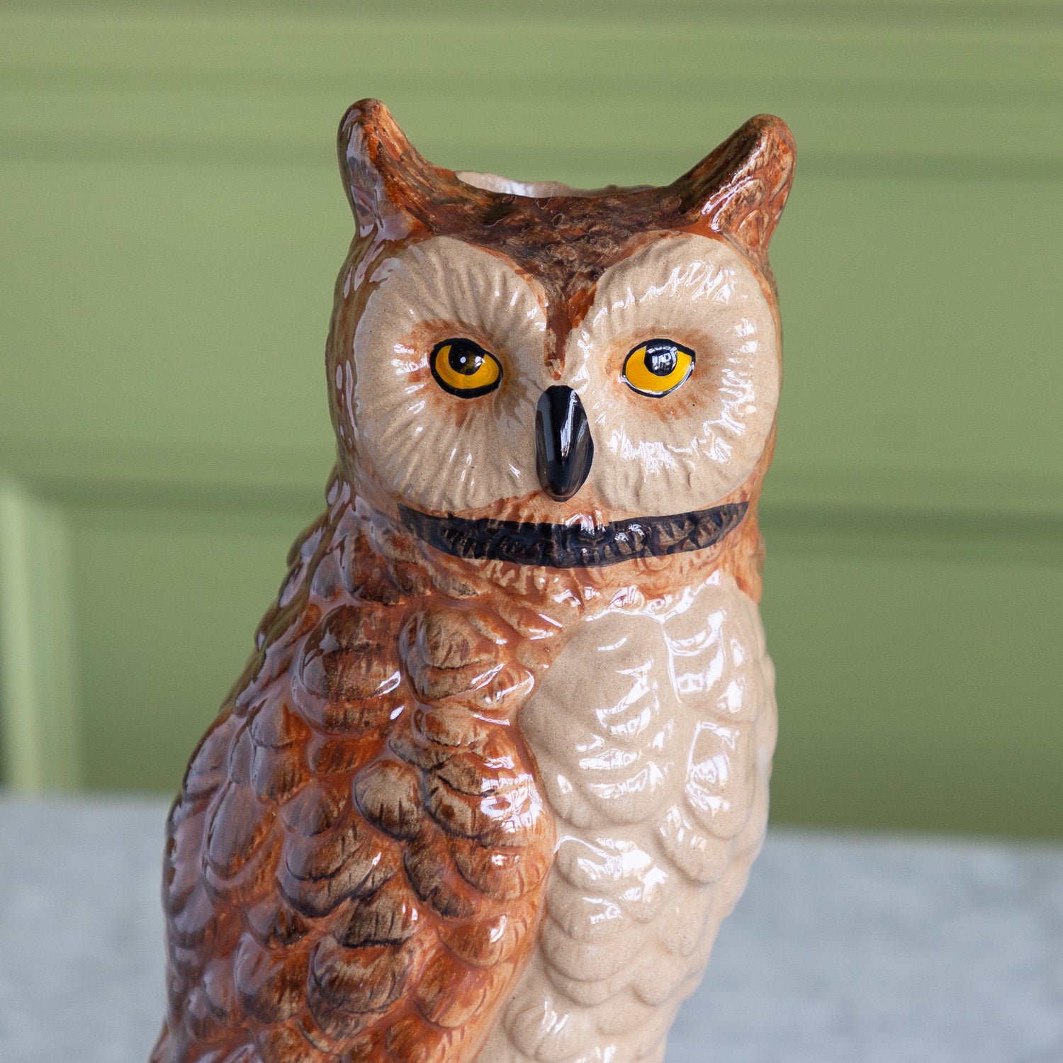 A ceramic owl figurine with detailed brown and beige feathers, yellow eyes, and a black beak, set against a soft green paneled background.
