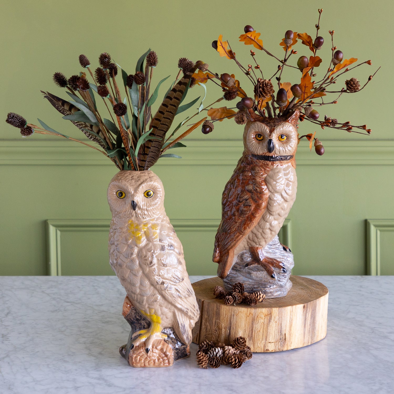 Two decorative owl figurines displayed on a light marble surface against a sage green paneled wall. One owl is cream-colored and stands on a branch-like base with dried leaves and pinecones, while the other is brown and stands on a round wood slice with autumn branches and acorns.