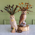 Two decorative owl figurines displayed on a light marble surface against a sage green paneled wall. One owl is cream-colored and stands on a branch-like base with dried leaves and pinecones, while the other is brown and stands on a round wood slice with autumn branches and acorns.