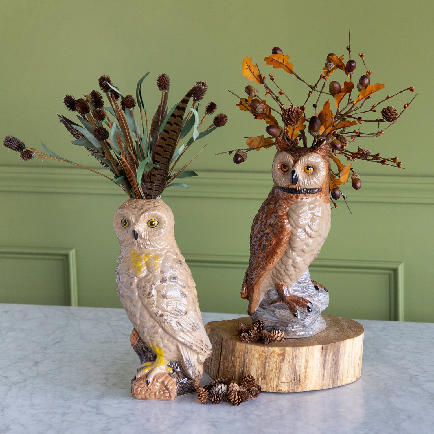 Two decorative owl figurines displayed on a light marble surface against a sage green paneled wall. The left owl is cream-colored with yellow accents and holds a bundle of brown feathers.