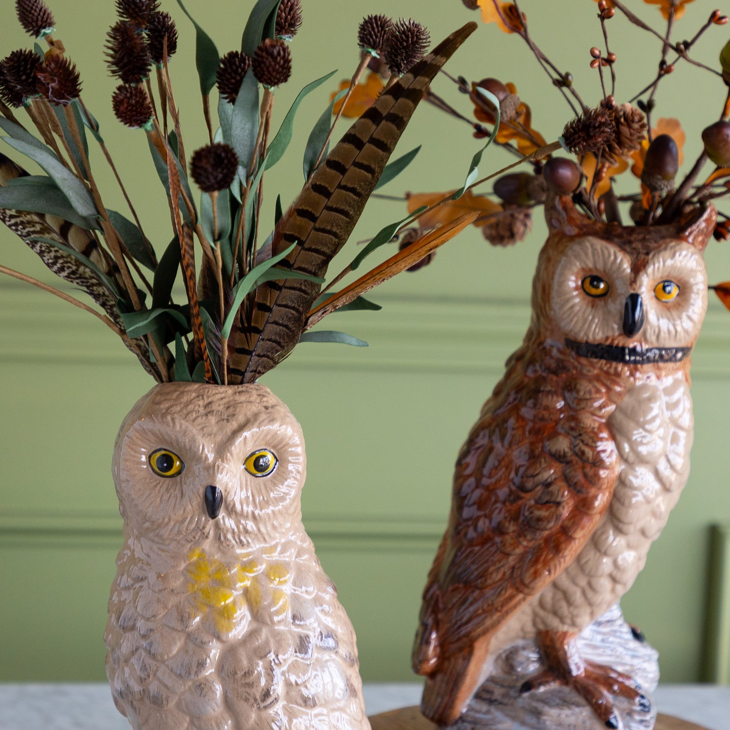 Two ceramic owl figurines with textured painted feathers, one in beige with yellow eyes and the other in brown, styled with dried branches and feathers in front of a green paneled wall.