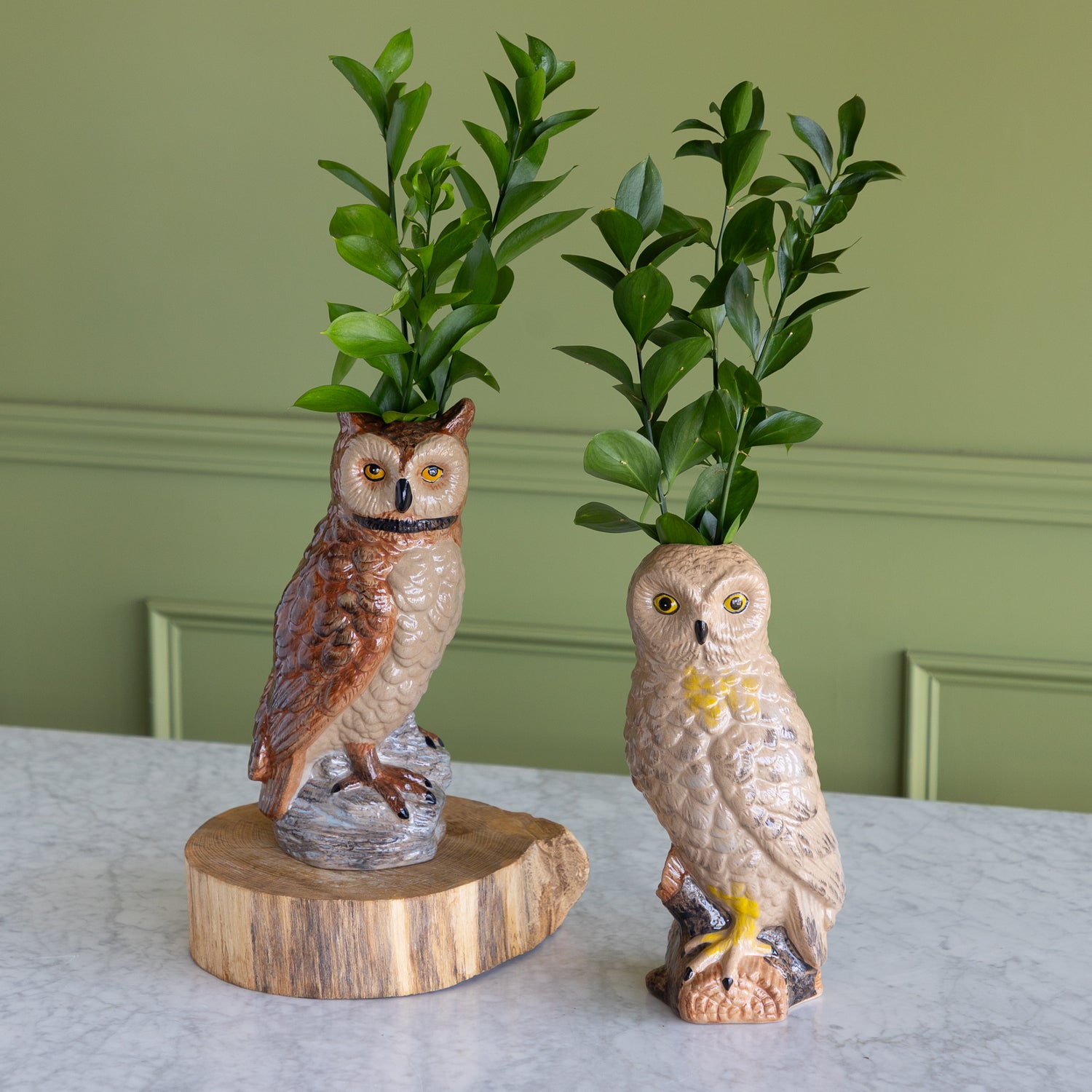 Two decorative owl figurines on a light marble surface against a sage green paneled wall. One owl is brown with a wood slice base and holds green leaves, while the other is beige with a small yellow accent and holds a small bird, both set against a styled interior backdrop.