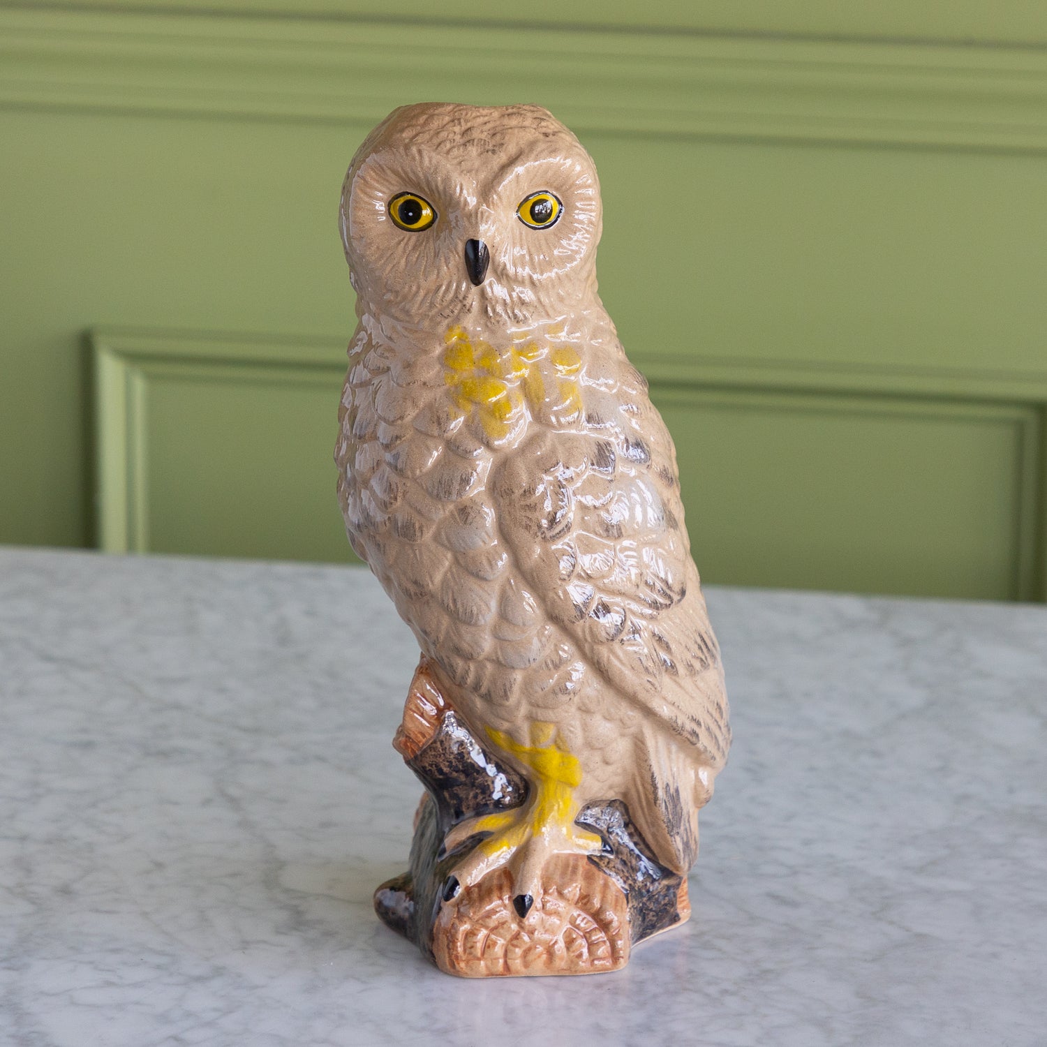 A ceramic owl figurine perched on a branch, featuring a textured beige and brown body with bright yellow accents around the eyes and chest, displayed on a marble surface against a green paneled wall.