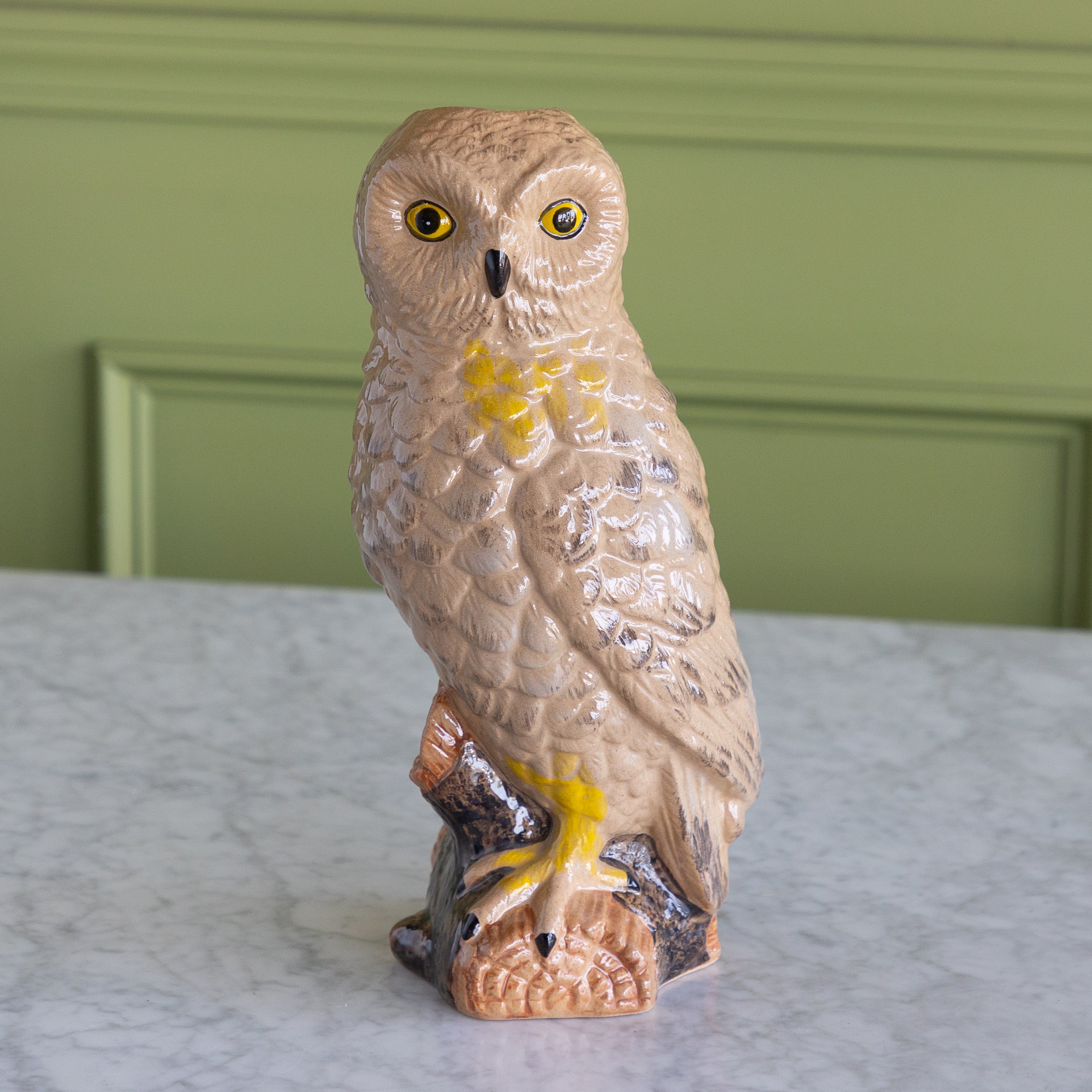 A ceramic owl figurine perched on a branch, featuring a textured beige and brown body with bright yellow accents around the eyes and chest, displayed on a marble surface against a green paneled wall.