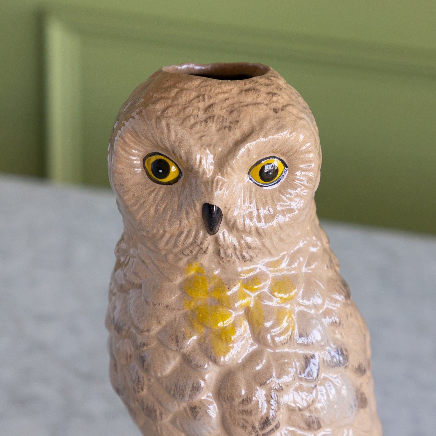A ceramic owl figurine with textured brown and beige feathers, bright yellow eyes, and a small black beak, shown close-up against a soft-focus green wall and light surface.
