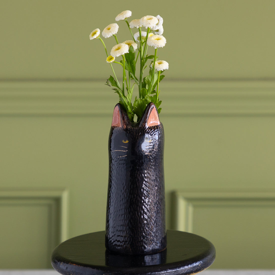 A glossy black cat-shaped ceramic vase with pink inner ears and gold whisker details, filled with white daisies, set on a round black pedestal against a sage green paneled wall.