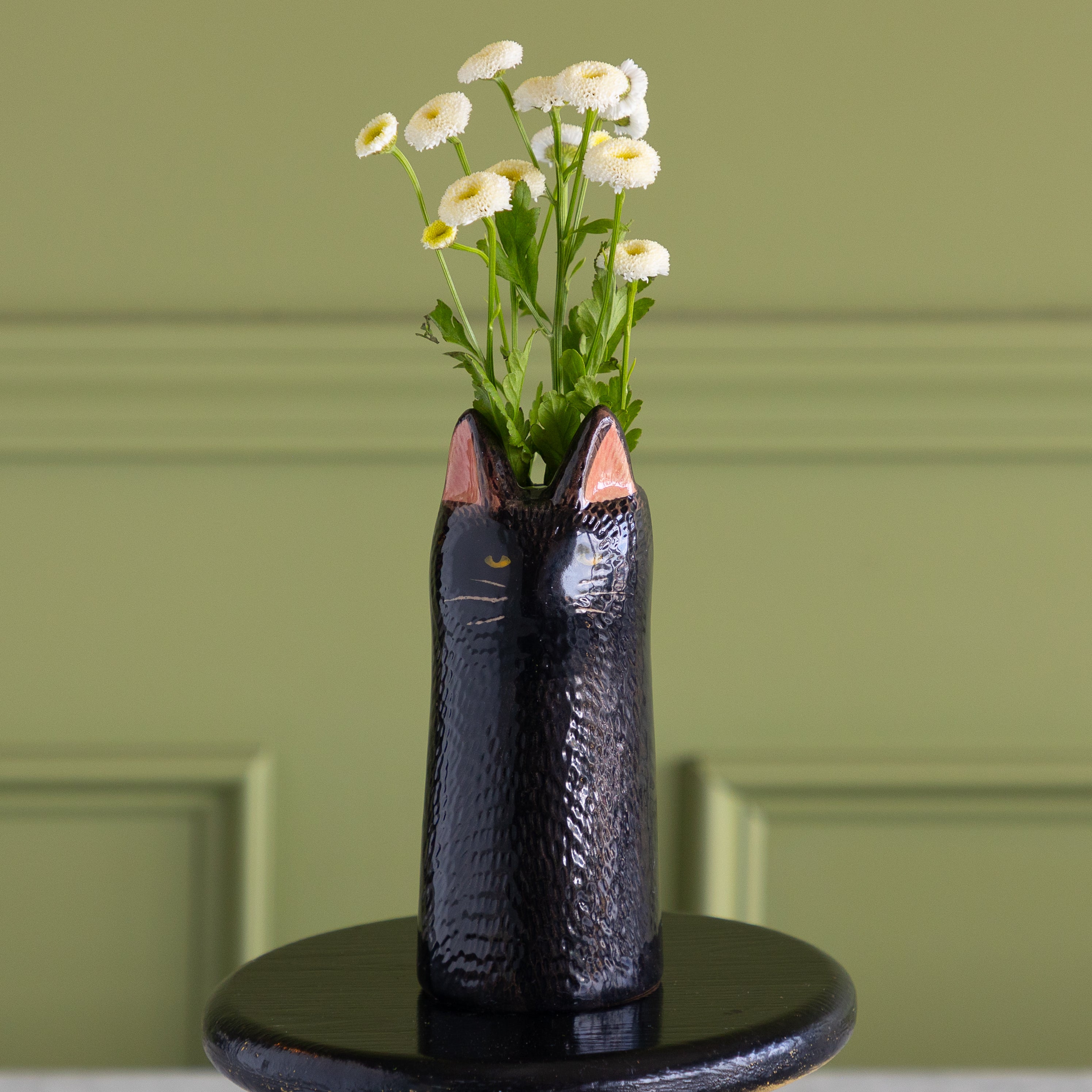 A glossy black cat-shaped ceramic vase with pink inner ears and gold whisker details, filled with white daisies, set on a round black pedestal against a sage green paneled wall.