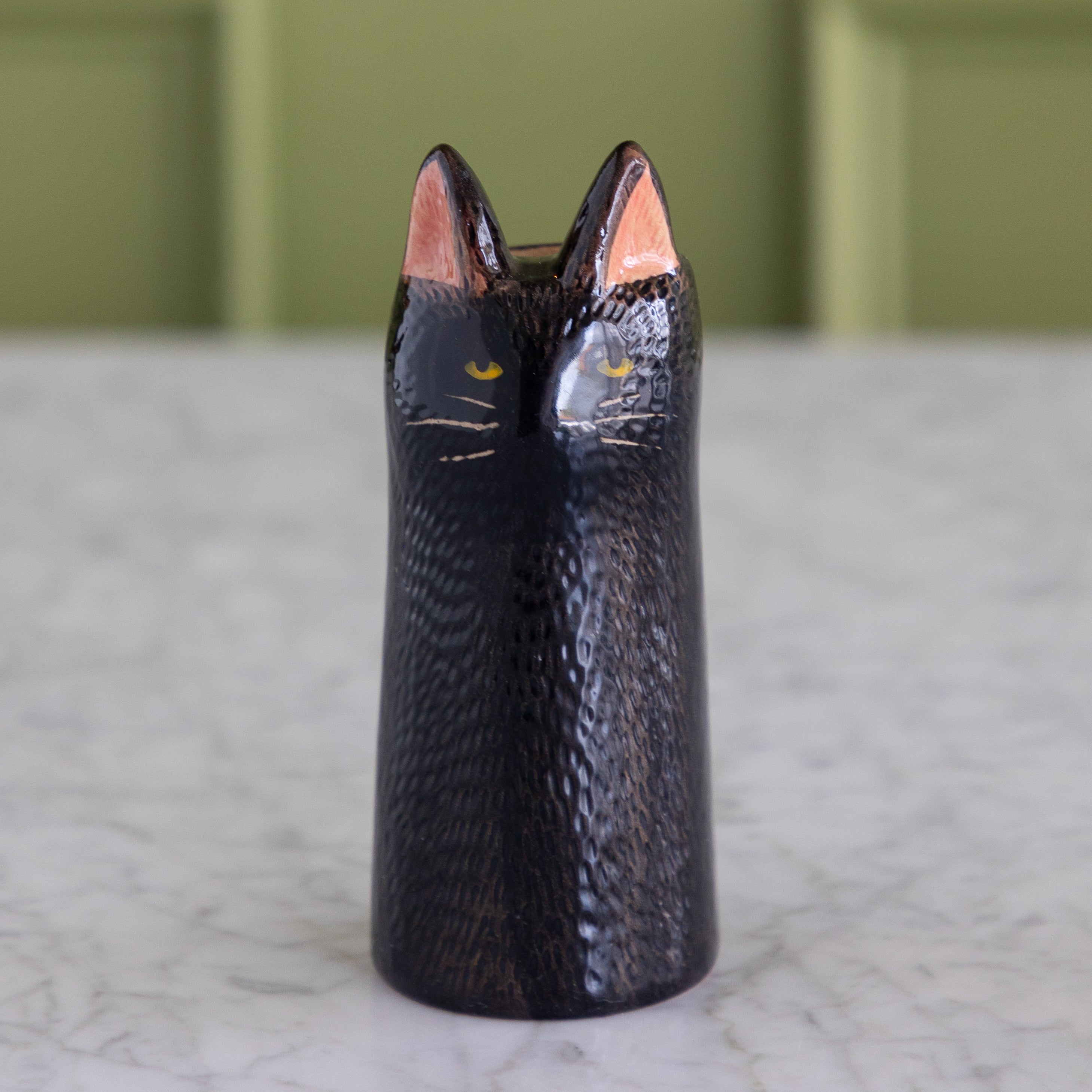 A glossy black cat-shaped vase with pointed ears, yellow eyes, and white whiskers, standing on a light marble surface with a soft-focus olive green background.