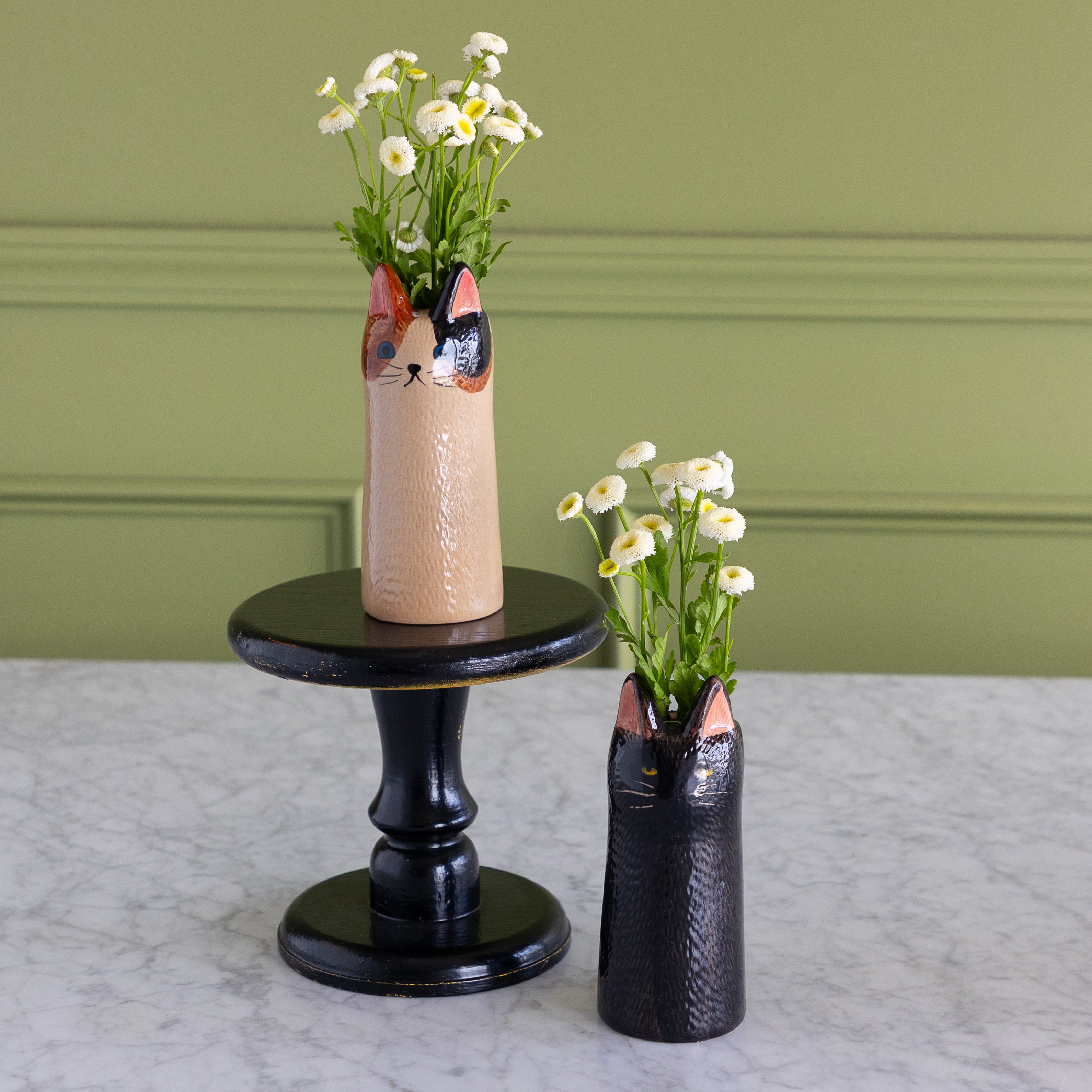 Two ceramic cat-shaped flower vases with textured glazes, one in beige and one in black, holding white daisies, displayed on a black pedestal stand and a white marble surface against a sage green paneled wall.