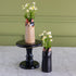 Two ceramic cat-shaped flower vases with textured glazes, one in beige and one in black, holding white daisies, displayed on a black pedestal stand and a white marble surface against a sage green paneled wall.