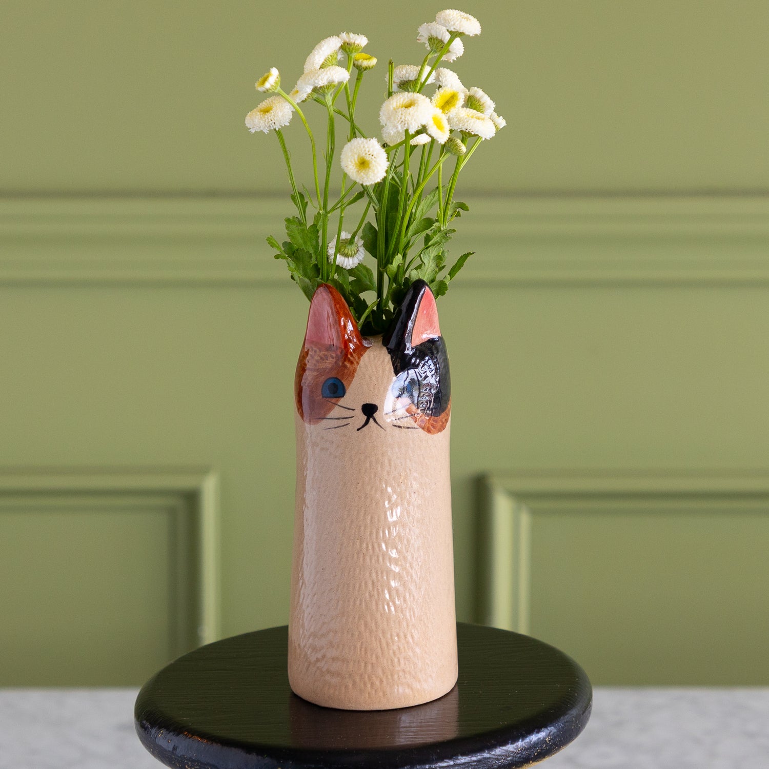 A cat-shaped ceramic vase filled with white daisies, displayed on a round black stand against a sage green paneled wall.