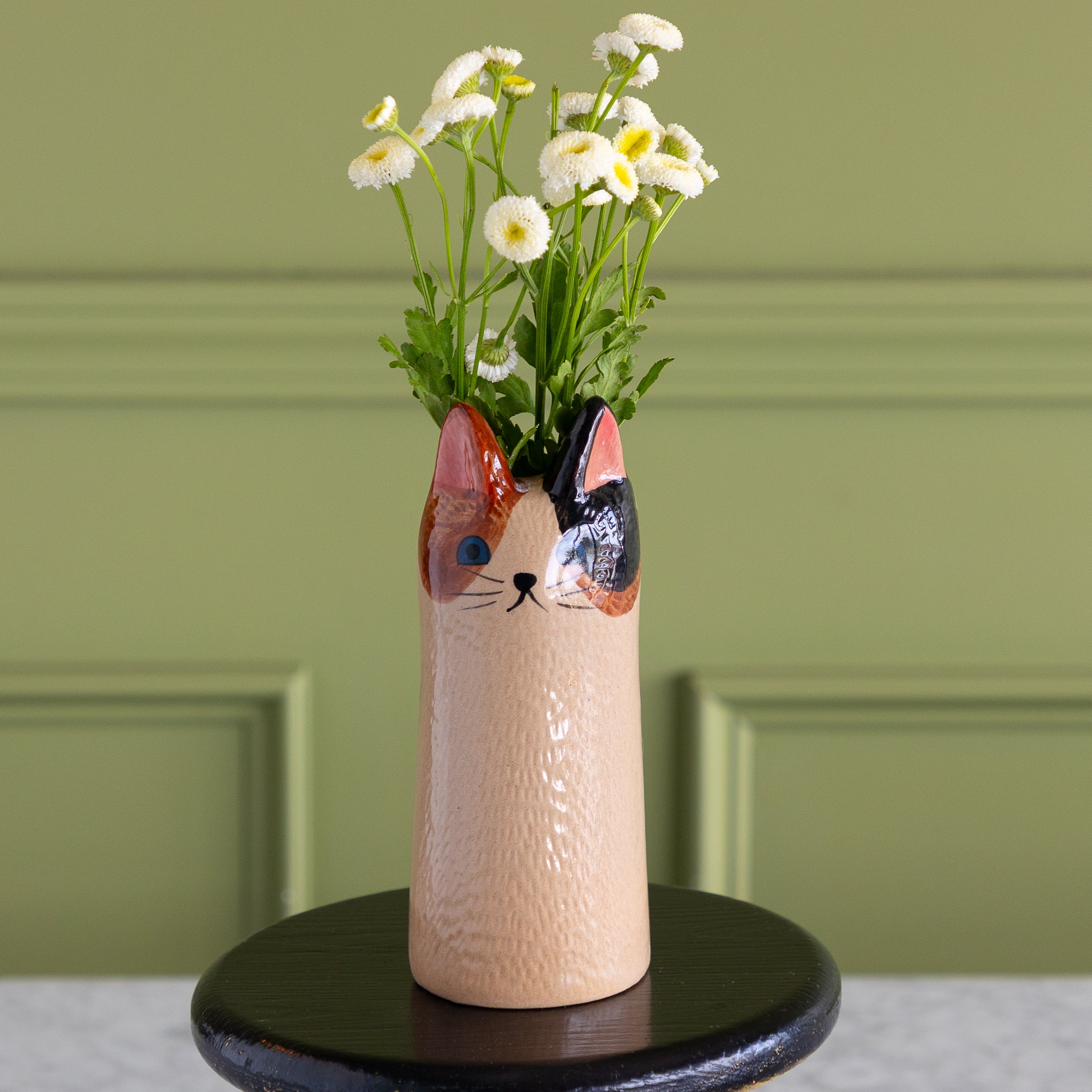 A cat-shaped ceramic vase filled with white daisies, displayed on a round black stand against a sage green paneled wall.