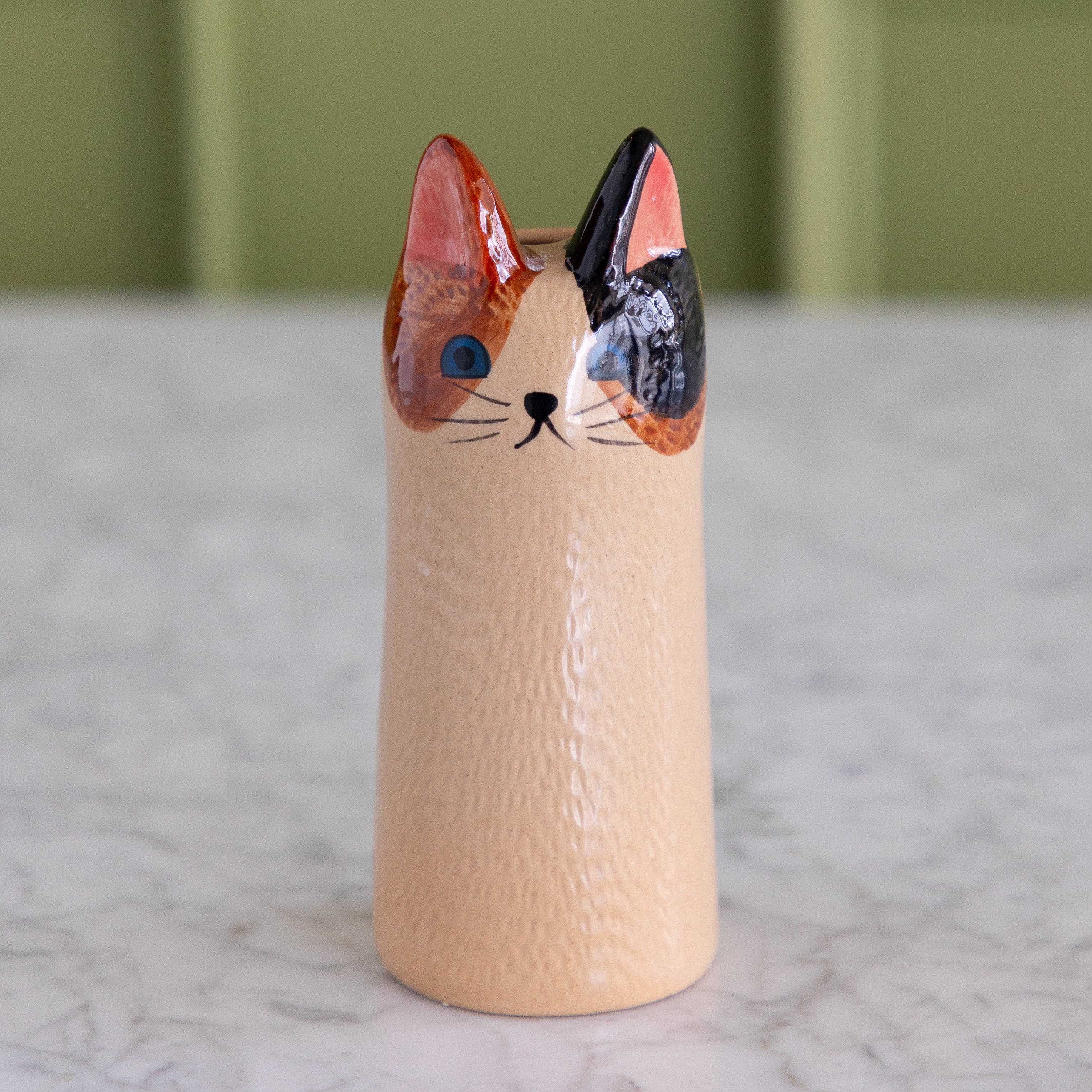 A small ceramic cat-shaped container or vase with a textured beige body, featuring a painted face with orange, black, pink, and blue details, standing on a light marble surface.