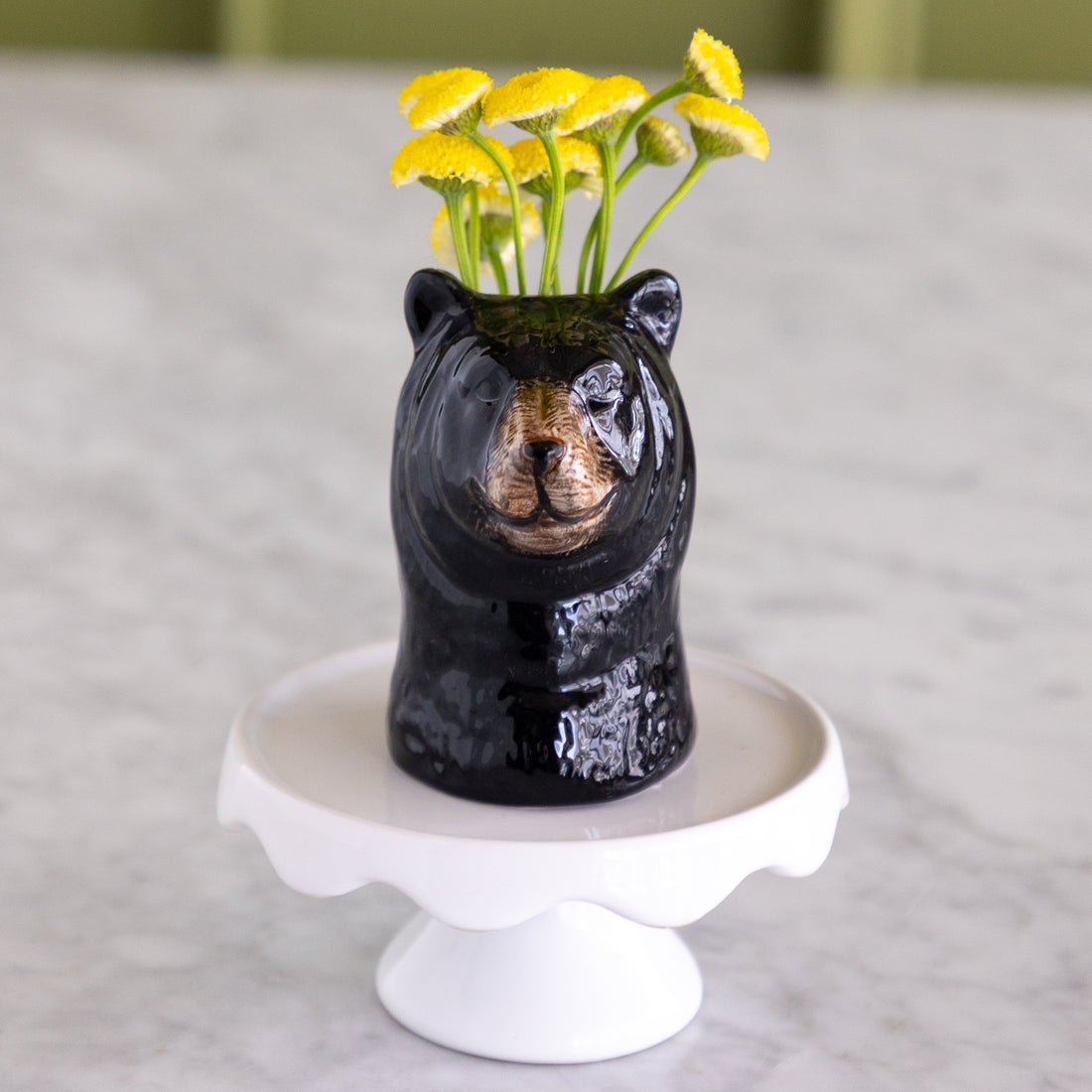 A glossy black bear-shaped ceramic vase filled with small yellow flowers, displayed on a white pedestal stand against a soft gray marble-like background.