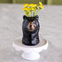 A glossy black bear-shaped ceramic vase filled with small yellow flowers, displayed on a white pedestal stand against a soft gray marble-like background.
