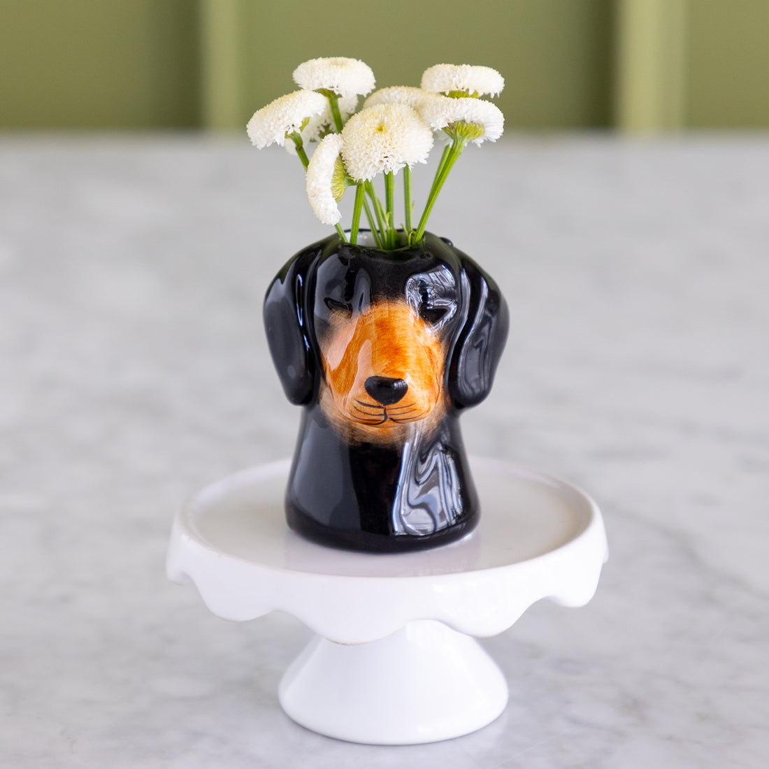 A glossy black ceramic dachshund-shaped vase filled with small white flowers, displayed on a white pedestal stand against a soft-focus indoor background.