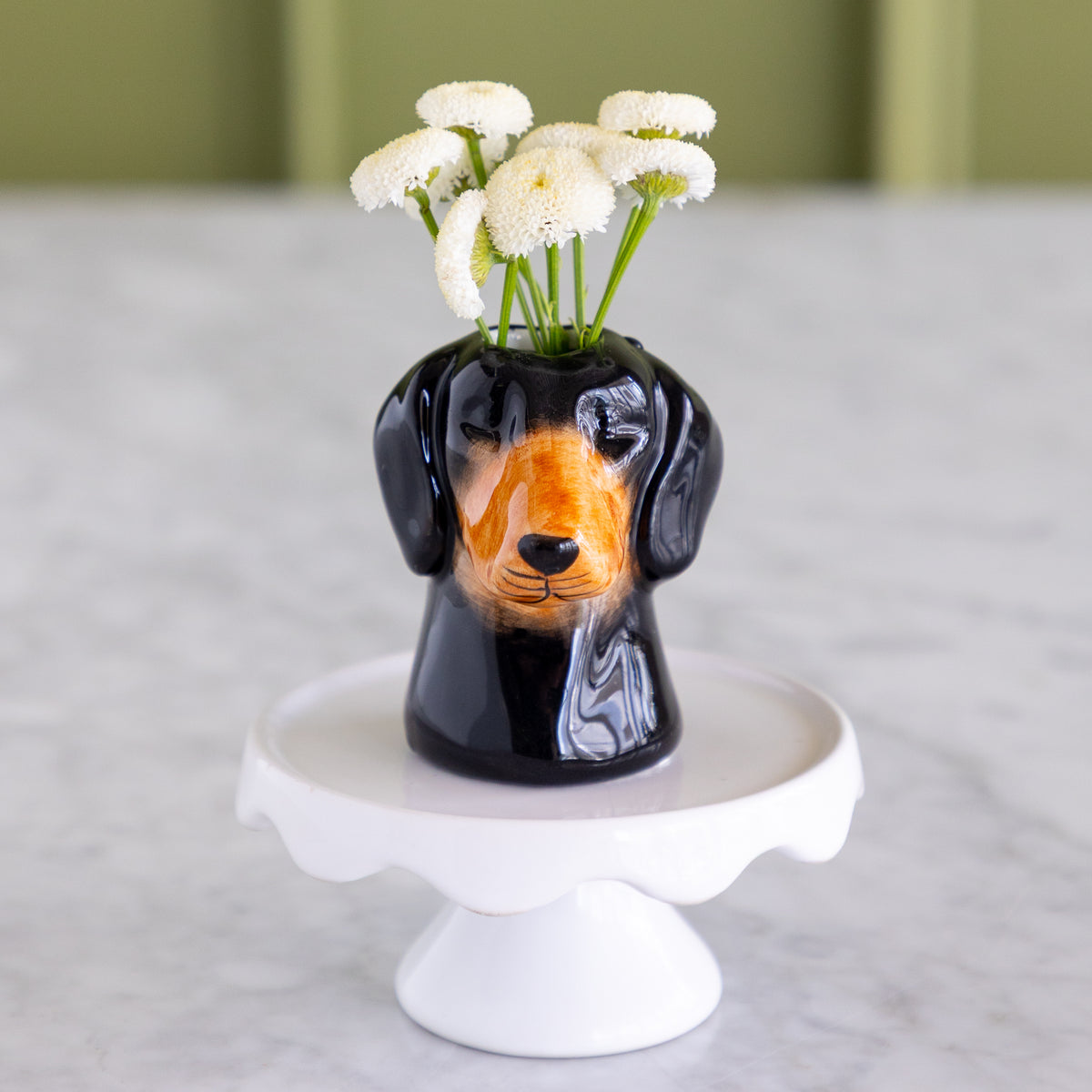 A glossy black ceramic dachshund-shaped vase filled with small white flowers, displayed on a white pedestal stand against a soft-focus indoor background.