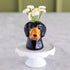 A glossy black ceramic dachshund-shaped vase filled with small white flowers, displayed on a white pedestal stand against a soft-focus indoor background.