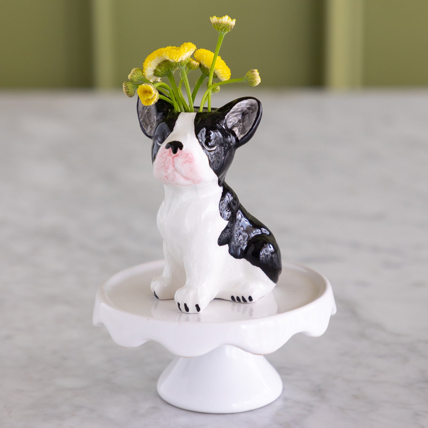 A ceramic French bulldog flower vase sitting on a white pedestal stand, with small yellow flowers placed in its raised head. The vase is photographed on a light marble-like surface with a softly blurred olive-green background.