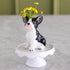 A ceramic French bulldog flower vase sitting on a white pedestal stand, with small yellow flowers placed in its raised head. The vase is photographed on a light marble-like surface with a softly blurred olive-green background.
