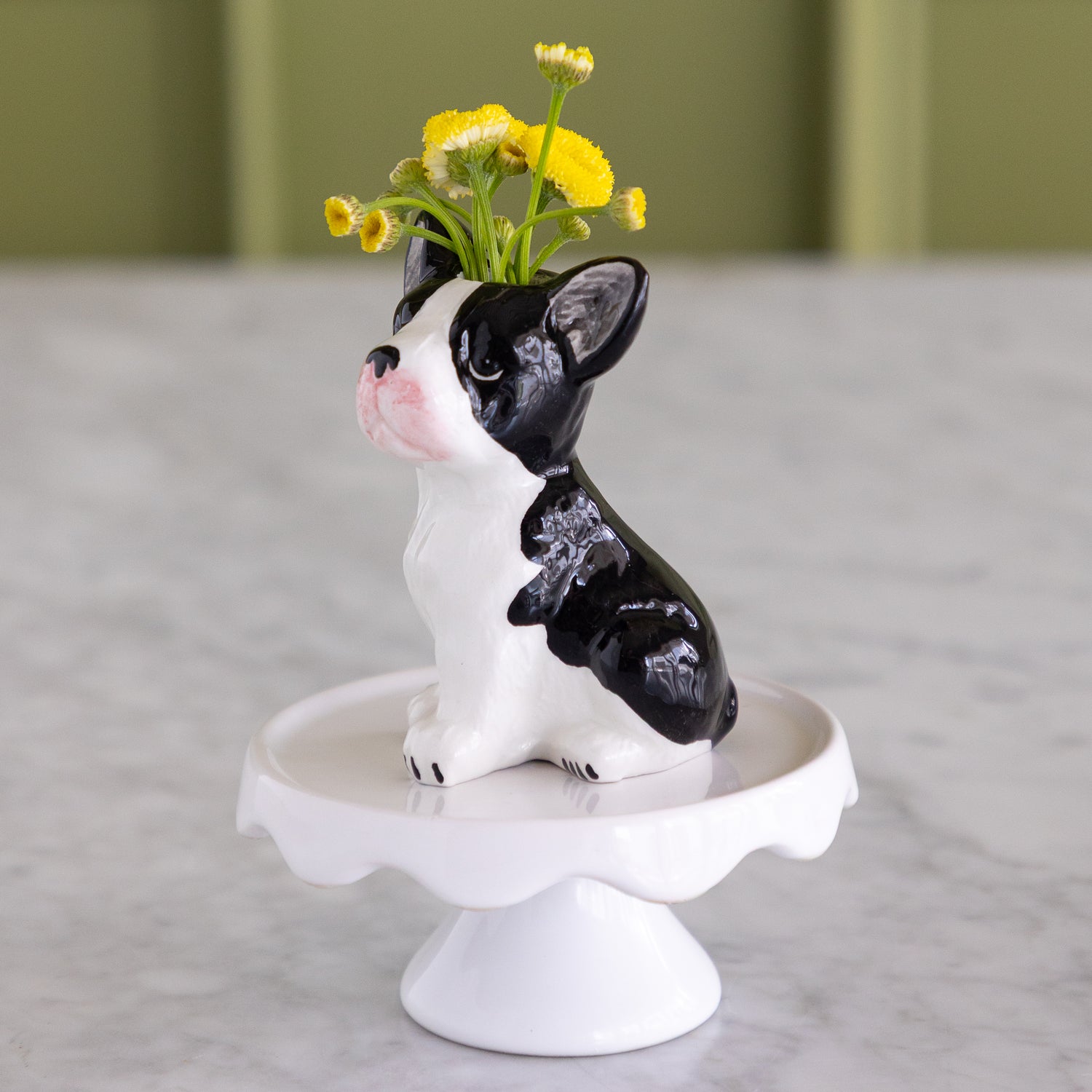 A black and white ceramic dog-shaped vase with yellow flowers in its mouth, sitting on a white pedestal stand on a marble surface.