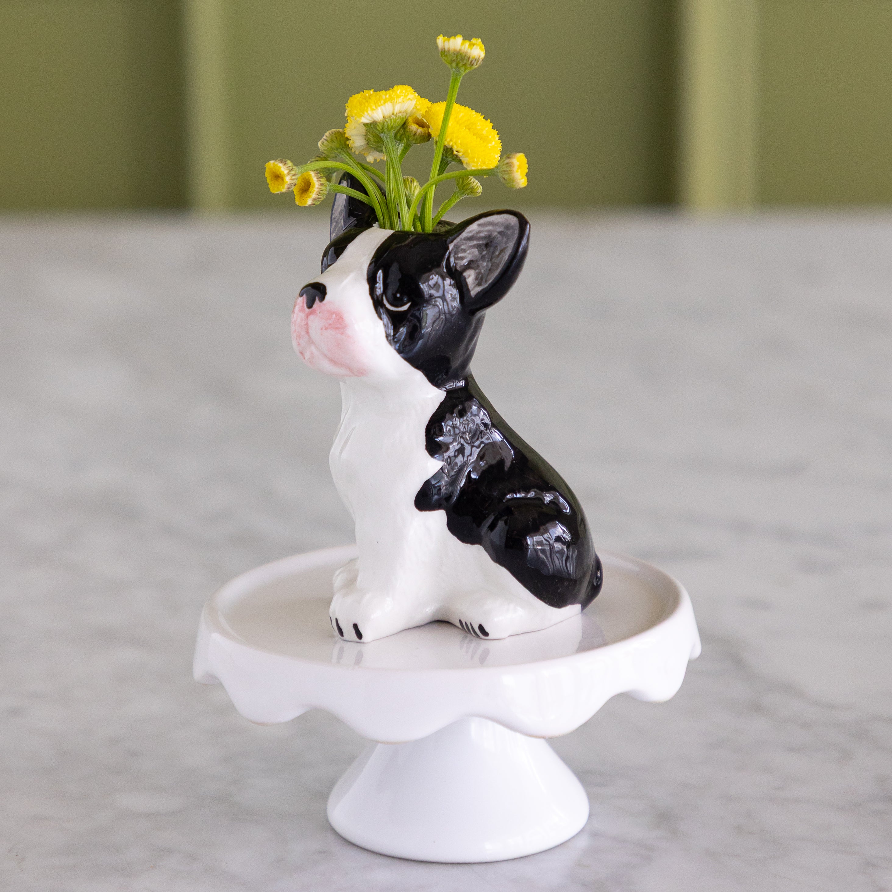 A black and white ceramic dog-shaped vase with yellow flowers in its mouth, sitting on a white pedestal stand on a marble surface.