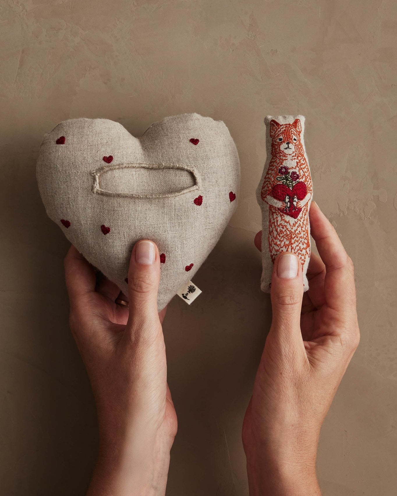 Heart-shaped fabric item with embroidered details held by hands against a beige background.