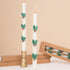 Two white taper candles decorated with green holly leaves and small red berries, shown with a gold candlestick holder and kraft gift boxes on a beige background.