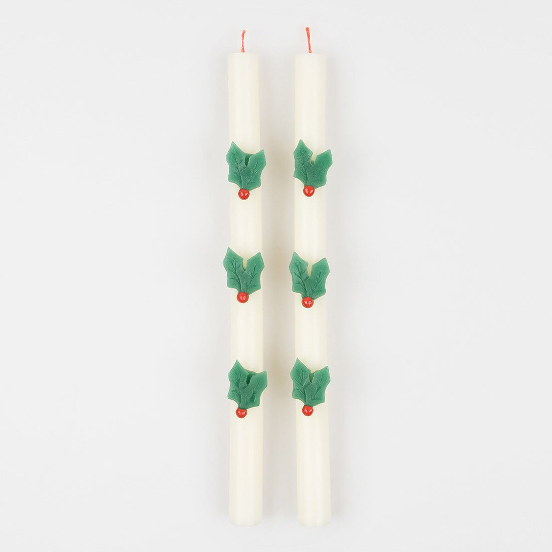 Two white taper candles decorated with green holly leaves and small red berries, shown upright on a plain light gray background.