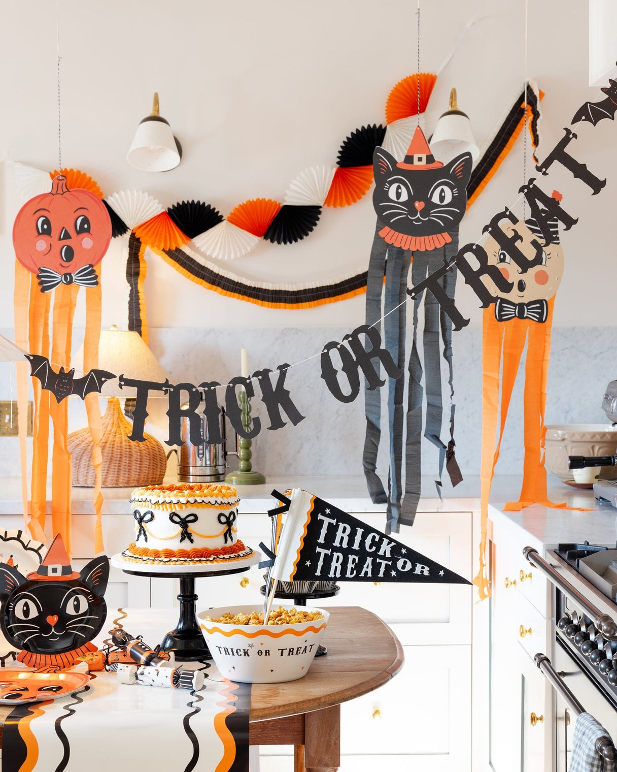 A Halloween-themed kitchen and dining area decorated with orange, black, and white paper fans, a black cat banner, a pumpkin, and a &
