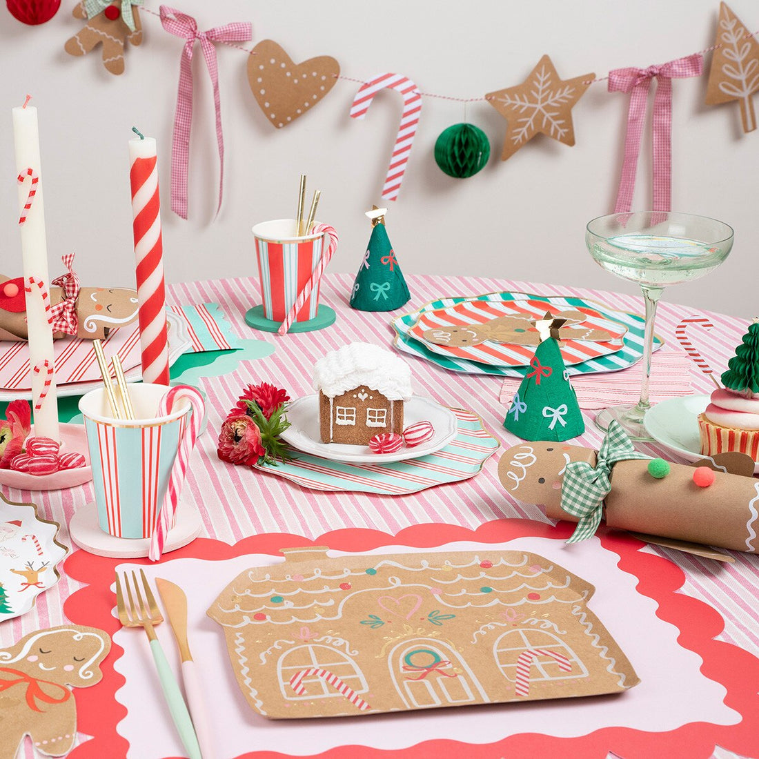 A festive Christmas gingerbread-themed party table setting with red and white striped tablecloth, gingerbread house placemats, candy cane cups, green party hats, and holiday decorations on a light wall.