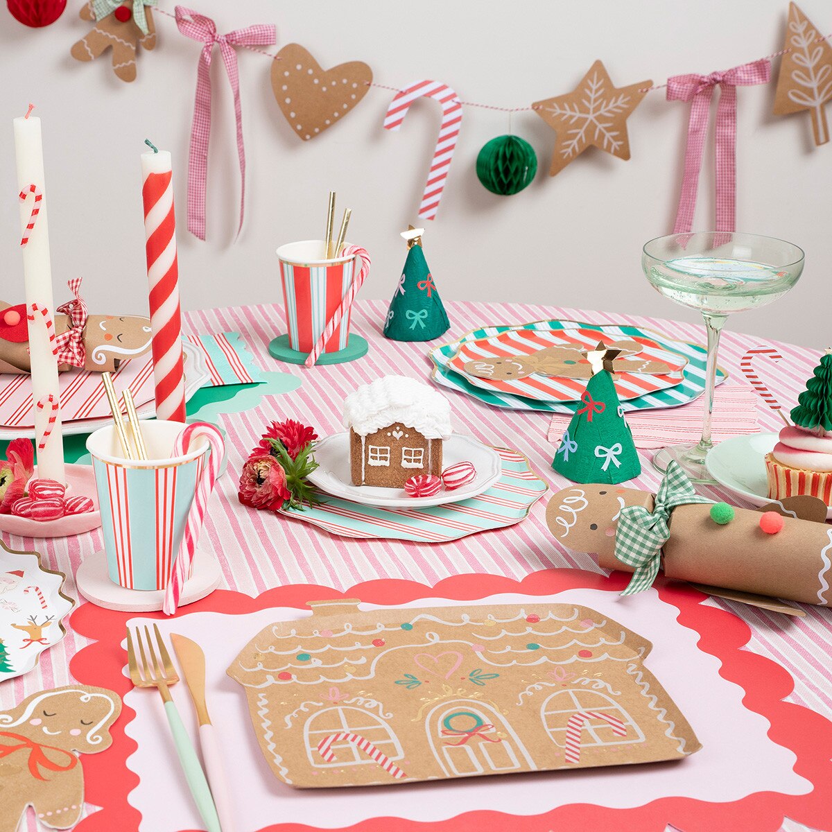 A festive Christmas gingerbread-themed party table setting with red and white striped tablecloth, gingerbread house placemats, candy cane cups, green party hats, and holiday decorations on a light wall.