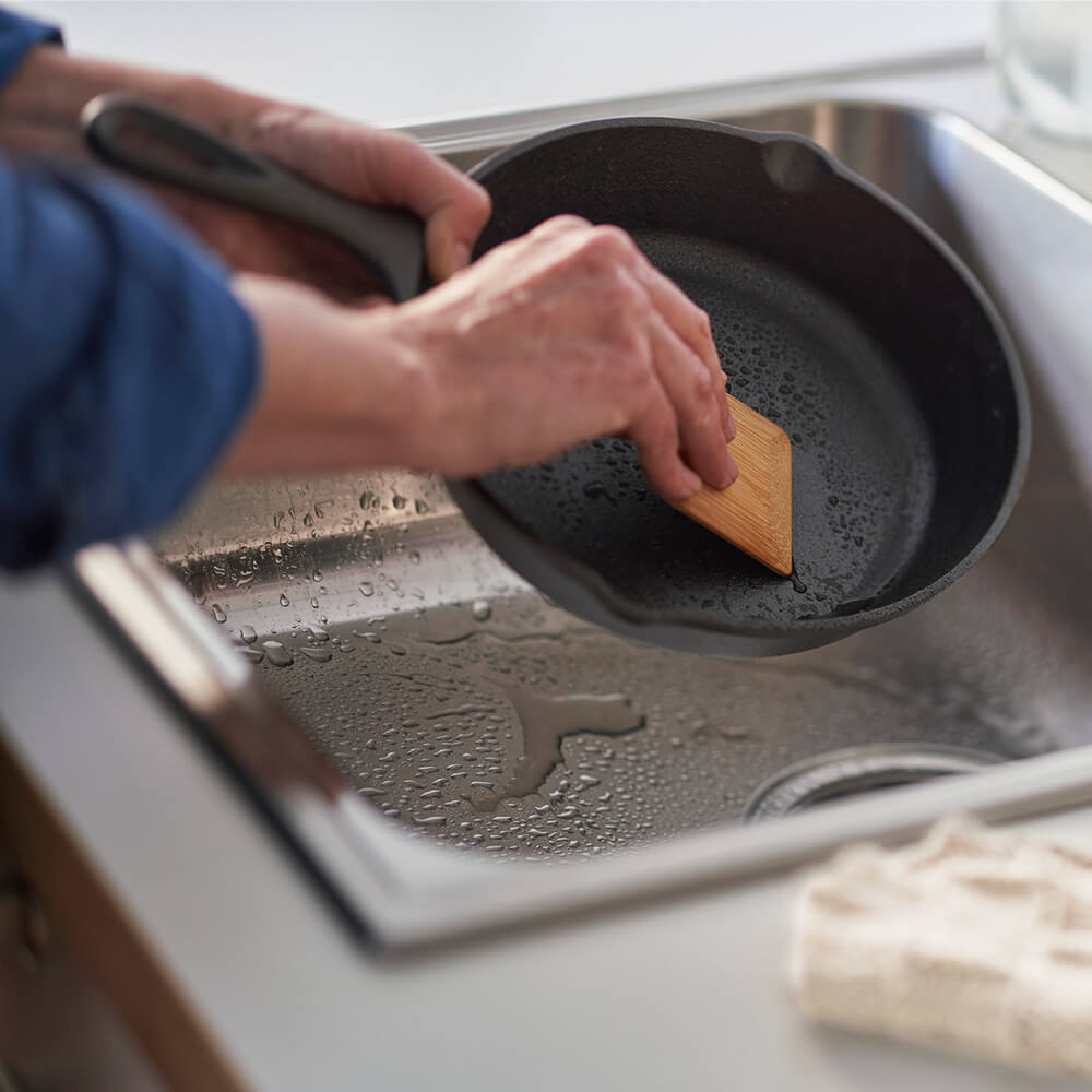 Person scrubbing a black frying pan in a sink with a bamboo scraper. 