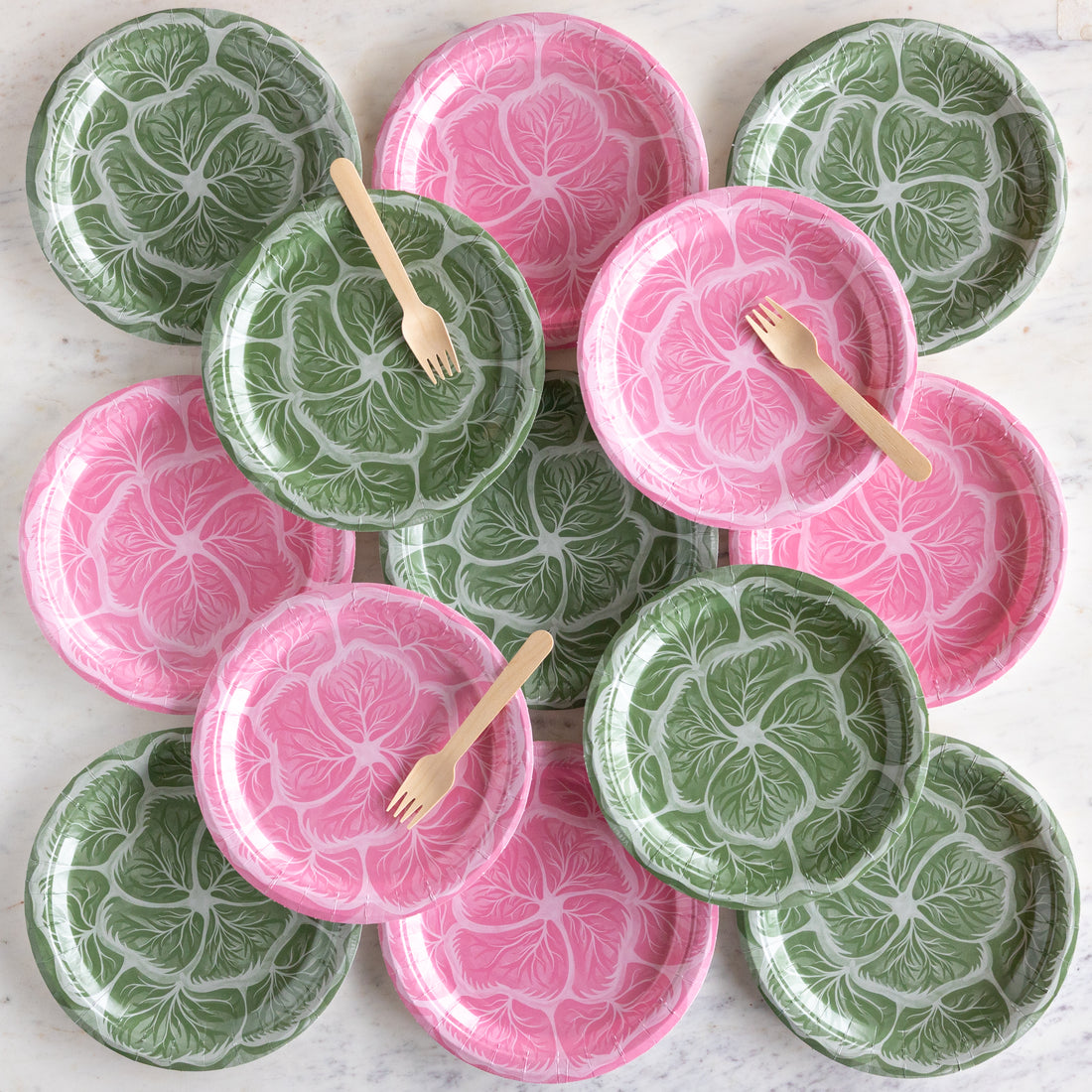 Set of pink and green cabbage patterned paper plates with wooden forks on a white surface.