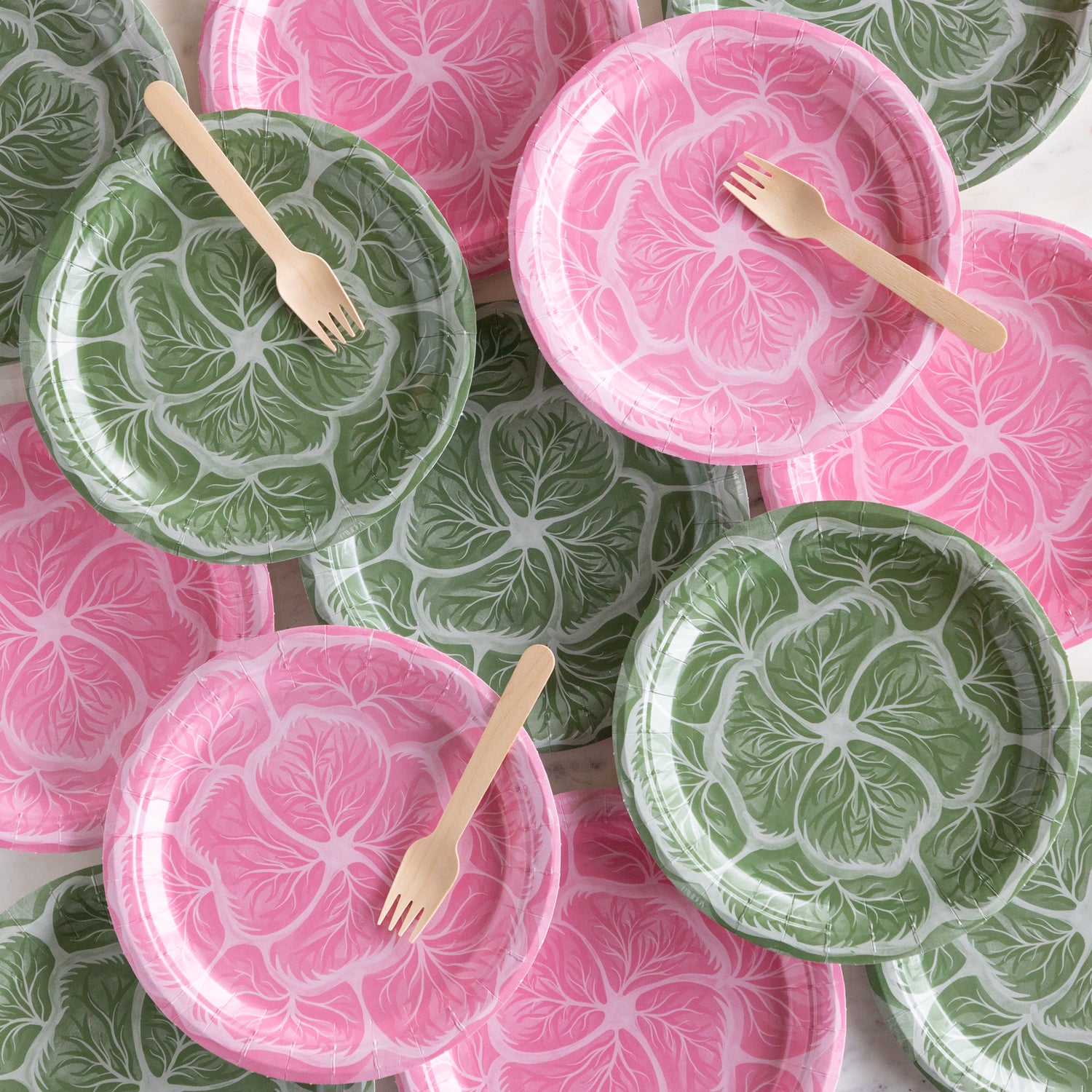 Set of pink and green cabbage patterned paper plates with wooden forks on a white surface.