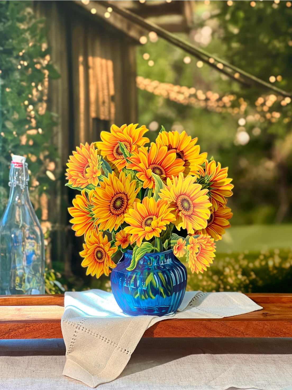 Bouquet of paper sunflowers in a blue vase on a wooden table outdoors.