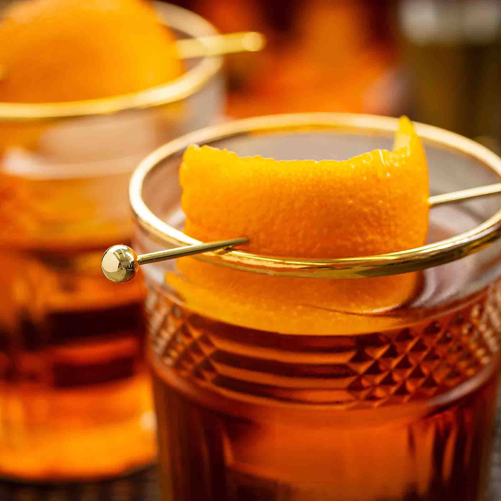 Close-up of a cocktail with a Gold Cocktail Pick piercing an orange peel garnish in a glass.