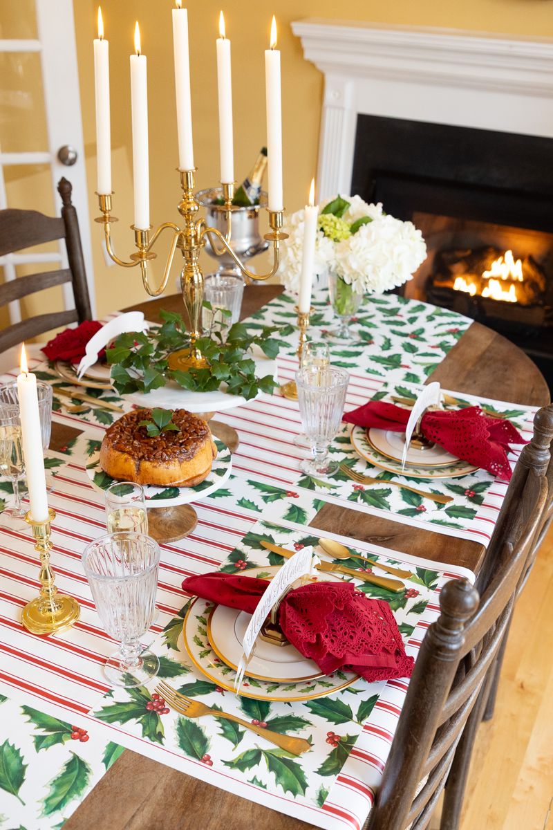 Dining table set for Christmas with holly leaf tablecloth, candles, and red napkins.