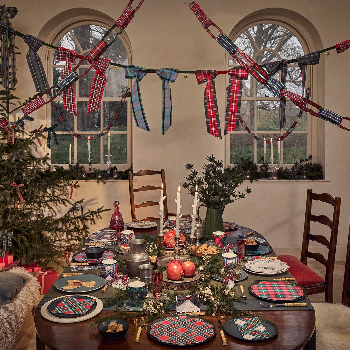 A festive holiday dining room decorated with red, green, and blue tartan plaid bunting garlands strung across two arched windows, a decorated Christmas tree, and a table set with matching plaid plates, napkins, and table runners.
