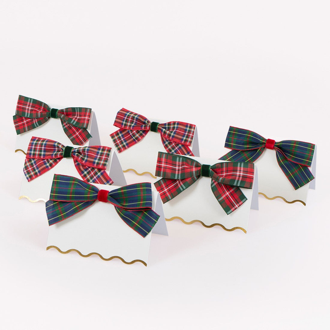 Six white paper place cards with wavy gold borders, each decorated with a ribbon bow in a tartan plaid pattern. The bows feature red, green, blue, and yellow plaid designs, with one bow in navy blue and red plaid and another in red and green plaid.